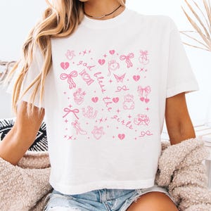 May include: White t-shirt with a pink graphic design featuring bows, hearts, and the text "God Bless the Girls." The shirt is a casual, short-sleeved style.