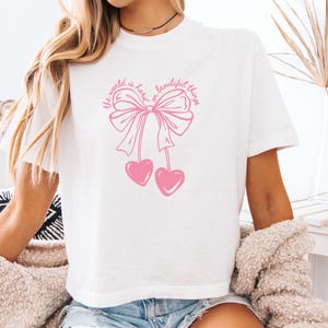May include: White t-shirt with a pink graphic of a bow and two hearts. The text "the world is has a beautiful thing" is above the bow. The shirt is short-sleeved.
