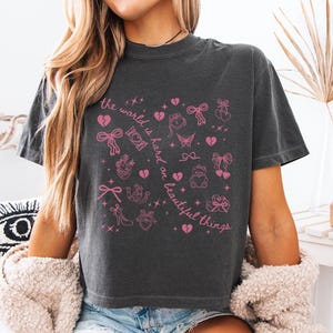 May include: Dark gray t-shirt with pink graphic illustrations and text. The text reads "the world is hard on beautiful things". The graphic includes hearts, bows, and other decorative elements. The shirt is a casual style.