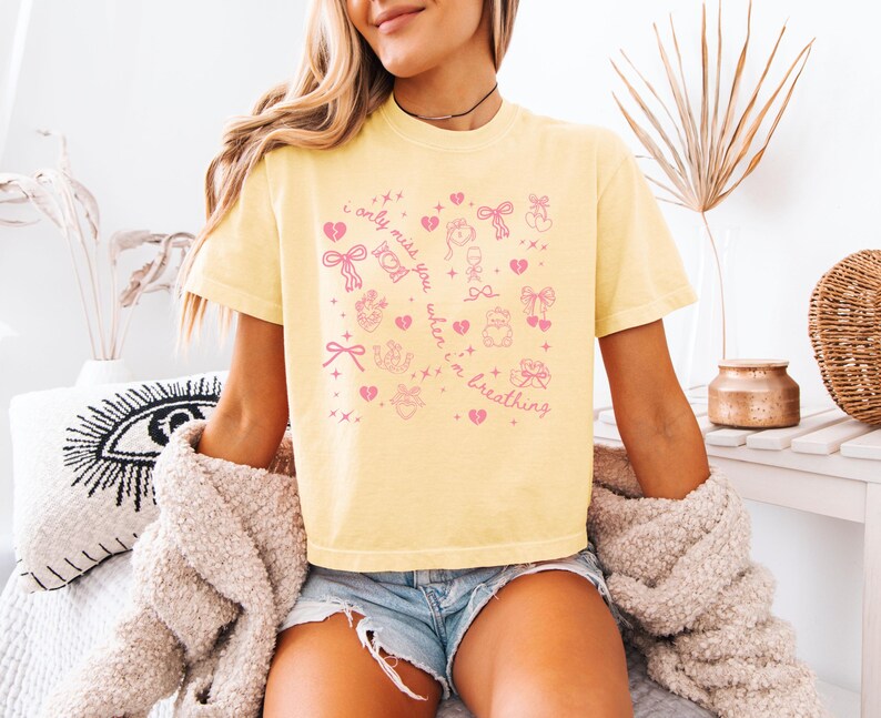 Pode incluir: Camiseta amarelo claro com ilustra&ccedil;&otilde;es gr&aacute;ficas rosa e o texto "I only miss you when I'm breathing". O design inclui cora&ccedil;&otilde;es, la&ccedil;os e outros s&iacute;mbolos fofos. A camiseta &eacute; um estilo casual e confort&aacute;vel.