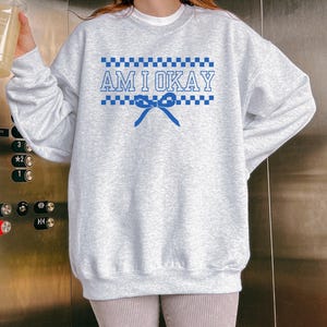 May include: A light gray sweatshirt with long sleeves. The front features the text "AM I OKAY" in blue, with a checkered pattern above and below the text, and a blue bow. The sweatshirt is a casual style.