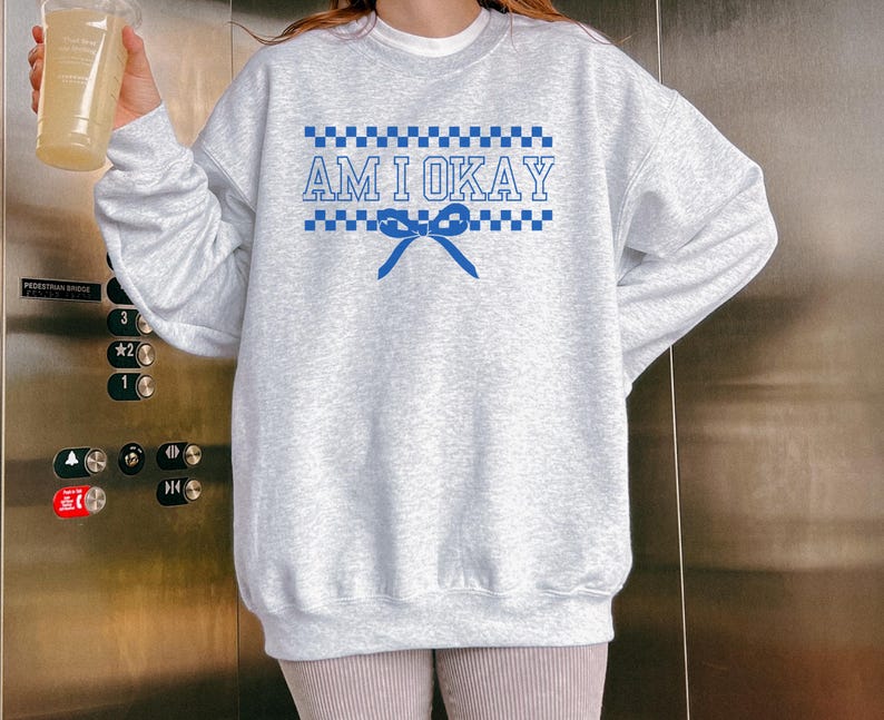 May include: A light gray sweatshirt with long sleeves. The front features the text "AM I OKAY" in blue, with a checkered pattern above and below the text, and a blue bow. The sweatshirt is a casual style.