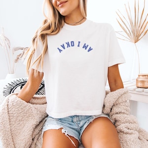 May include: White t-shirt with the text "AM I OKAY" in blue arched across the chest. The shirt is paired with denim shorts. The model is wearing a beige sweater and a black necklace. The background includes decorative items.