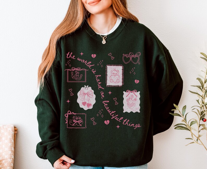 K&ouml;nnte beinhalten: Dunkelgr&uuml;nes Sweatshirt mit rosa Grafikaufn&auml;hern und dem Text "the world is hard on beautiful things". Die Aufn&auml;her zeigen Herzen, Schleifen und einen Teddyb&auml;ren. Das Sweatshirt hat einen Rundhalsausschnitt und lange &Auml;rmel.