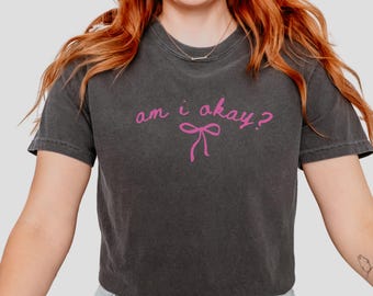 May include: Dark gray t-shirt with the phrase "am i okay?" in pink script, featuring a bow graphic. The shirt has a crew neck and short sleeves. The overall style is casual and comfortable.