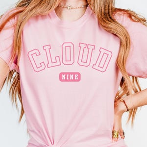 May include: A light pink t-shirt with the words "CLOUD NINE" in a pink collegiate font. The word "NINE" is in a smaller pink rectangle below the word "CLOUD". The shirt is made of a soft material.