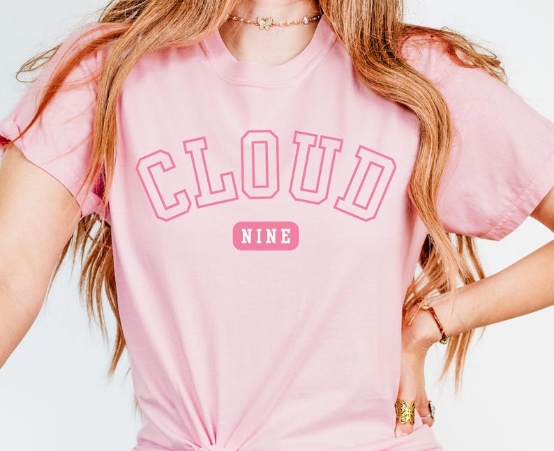 May include: A light pink t-shirt with the words "CLOUD NINE" in a pink collegiate font. The word "NINE" is in a smaller pink rectangle below the word "CLOUD". The shirt is made of a soft material.