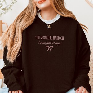 Może przedstawiać: Czarna bluza z r&oacute;żowym napisem "THE WORLD IS HARD ON beautiful things" i kokardą. Tekst jest napisany kursywą. Bluza ma długie rękawy i okrągły dekolt.