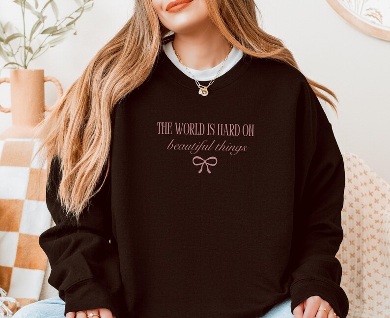 Może przedstawiać: Czarna bluza z r&oacute;żowym napisem "THE WORLD IS HARD ON beautiful things" i kokardą. Tekst jest napisany kursywą. Bluza ma długie rękawy i okrągły dekolt.