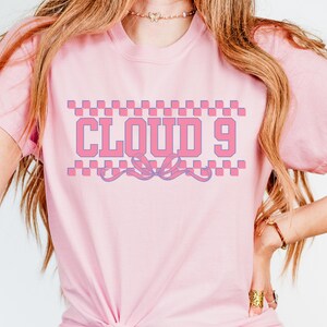 Puede incluir: Camiseta rosa claro con el texto "CLOUD 9" en dise&ntilde;o rosa y morado. El texto est&aacute; enmarcado por un patr&oacute;n de cuadros y una cinta decorativa. La camiseta tiene mangas cortas.