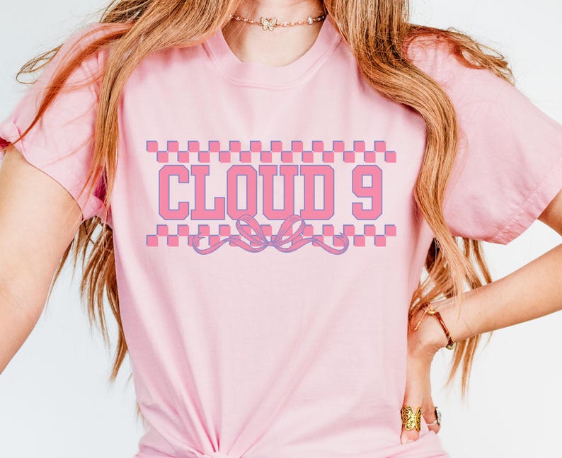 Puede incluir: Camiseta rosa claro con el texto "CLOUD 9" en dise&ntilde;o rosa y morado. El texto est&aacute; enmarcado por un patr&oacute;n de cuadros y una cinta decorativa. La camiseta tiene mangas cortas.