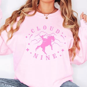 K&ouml;nnte beinhalten: Ein hellrosa Sweatshirt mit Rundhalsausschnitt. Auf der Vorderseite befindet sich eine rosa Grafik eines Pferdes und Reiters, umgeben von Wolken und Sternen, mit den Worten "CLOUD NINE" in einem kreisf&ouml;rmigen Design.