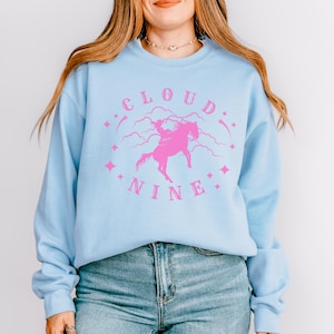 K&ouml;nnte beinhalten: Hellblaues Sweatshirt mit einem rosa Aufdruck mit der Aufschrift "CLOUD NINE" mit einem Einhorn und Sternen. Das Sweatshirt hat lange &Auml;rmel und einen Rundhalsausschnitt. Das Model tr&auml;gt eine blaue Jeans.