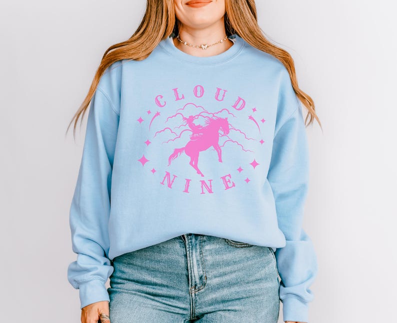 K&ouml;nnte beinhalten: Hellblaues Sweatshirt mit einem rosa Aufdruck mit der Aufschrift "CLOUD NINE" mit einem Einhorn und Sternen. Das Sweatshirt hat lange &Auml;rmel und einen Rundhalsausschnitt. Das Model tr&auml;gt eine blaue Jeans.