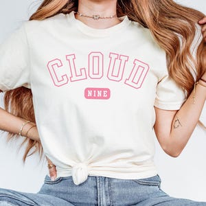 May include: Cream-colored t-shirt with the words "CLOUD NINE" in pink block letters. The word "NINE" is on a pink rectangular background. The shirt is a casual, short-sleeved style.