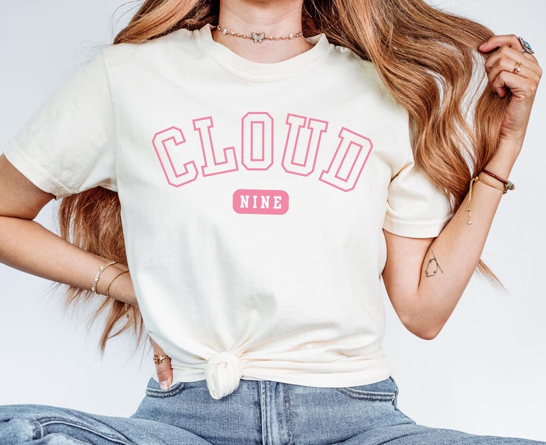 May include: Cream-colored t-shirt with the words "CLOUD NINE" in pink block letters. The word "NINE" is on a pink rectangular background. The shirt is a casual, short-sleeved style.