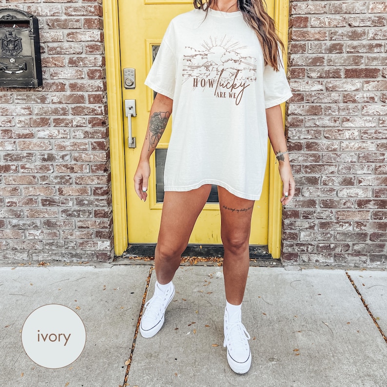 May include: A woman wearing a white oversized t-shirt with the text "How lucky are we" printed on it. The shirt has a brown graphic of a sunset with clouds. She is wearing white sneakers and standing in front of a yellow door.