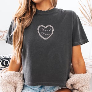 May include: Dark gray t-shirt with a crew neck. The shirt features a heart-shaped design made of small white beads, with the words "Cloud Nine" written inside the heart in a cursive font. The shirt is made of a soft material.