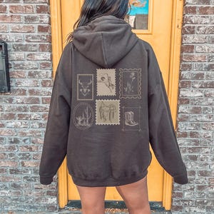 May include: A dark brown hoodie featuring several vintage-style postage stamp designs on the back. The stamps depict a bull, a cowboy, a flower, a cactus, a horse and a cowboy boot. The hoodie has a hood and a kangaroo pocket.