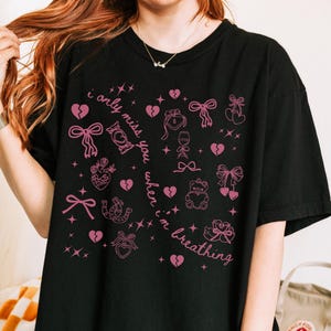 May include: Black t-shirt with a pink graphic design featuring hearts, bows, and the text "i only miss you when i'm breathing." The design includes various illustrations and stars. A casual, comfortable tee.