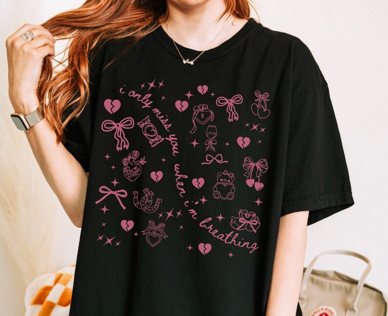 May include: Black t-shirt with a pink graphic design featuring hearts, bows, and the text "i only miss you when i'm breathing." The design includes various illustrations and stars. A casual, comfortable tee.