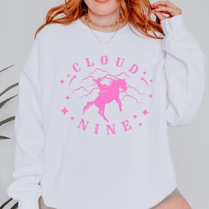 K&ouml;nnte beinhalten: Wei&szlig;es Sweatshirt mit einem rosa Aufdruck. Der Aufdruck zeigt die Worte "CLOUD NINE" in einem kreisf&ouml;rmigen Design, mit einer rosa Illustration eines Pferdes und Reiters in der Mitte. Rosa Sterne und Wolken vervollst&auml;ndigen das Design.