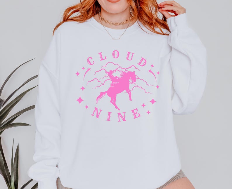 K&ouml;nnte beinhalten: Wei&szlig;es Sweatshirt mit einem rosa Aufdruck. Der Aufdruck zeigt die Worte "CLOUD NINE" in einem kreisf&ouml;rmigen Design, mit einer rosa Illustration eines Pferdes und Reiters in der Mitte. Rosa Sterne und Wolken vervollst&auml;ndigen das Design.