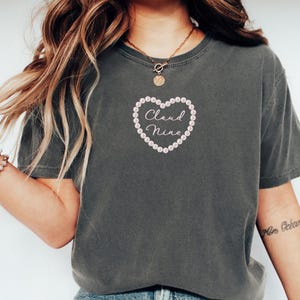 May include: Dark gray t-shirt with a heart-shaped design. The heart is outlined with small, light pink beads and contains the words "Cloud Nine" in a cursive font. The shirt has short sleeves and a crew neck.