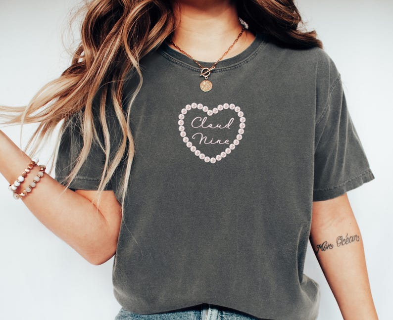 May include: Dark gray t-shirt with a heart-shaped design. The heart is outlined with small, light pink beads and contains the words "Cloud Nine" in a cursive font. The shirt has short sleeves and a crew neck.