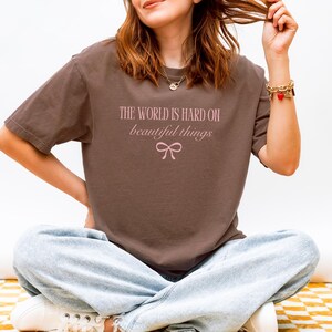 Puede incluir: Una camiseta marr&oacute;n con el texto "THE WORLD IS HARD ON beautiful things" en rosa claro, con un lazo gr&aacute;fico. La camiseta se combina con vaqueros azul claro y zapatillas blancas.