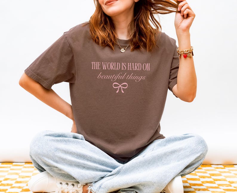 Puede incluir: Una camiseta marr&oacute;n con el texto "THE WORLD IS HARD ON beautiful things" en rosa claro, con un lazo gr&aacute;fico. La camiseta se combina con vaqueros azul claro y zapatillas blancas.
