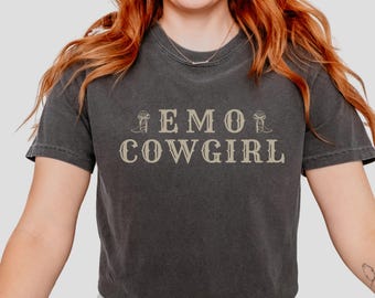 May include: A dark gray t-shirt with the text "EMO COWGIRL" in a vintage-style font. The text is surrounded by two small images of cowboy boots.