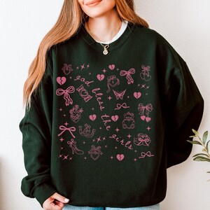 Może przedstawiać: Ciemnozielona bluza z białym kołnierzem, z r&oacute;żowym wzorem graficznym. Wz&oacute;r zawiera kokardy, serca, motyle i napis "god bless the girls". Swobodny, wygodny top.