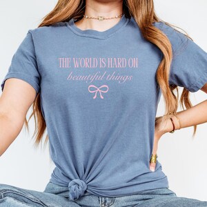 Puede incluir: Una camiseta azul polvoriento con el texto rosa "THE WORLD IS HARD ON beautiful things" y un gr&aacute;fico de lazo rosa. La camiseta est&aacute; anudada a la cintura.