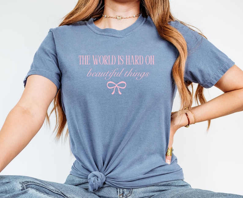 Puede incluir: Una camiseta azul polvoriento con el texto rosa "THE WORLD IS HARD ON beautiful things" y un gr&aacute;fico de lazo rosa. La camiseta est&aacute; anudada a la cintura.