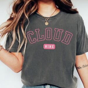May include: Dark gray t-shirt with the words "CLOUD NINE" in pink block letters. The word "NINE" is on a pink rectangular background. The shirt has short sleeves and a crew neck.