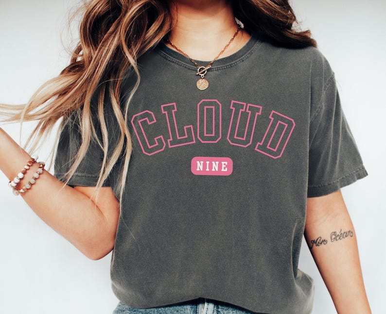 May include: Dark gray t-shirt with the words "CLOUD NINE" in pink block letters. The word "NINE" is on a pink rectangular background. The shirt has short sleeves and a crew neck.
