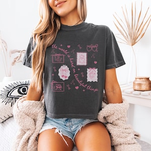 May include: Dark gray t-shirt with pink graphic illustrations of bows, stamps, and hearts. The text "the world is hard on beautiful things" is printed in pink. The shirt is a casual, comfortable style.