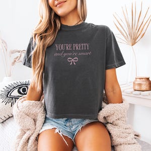 May include: A charcoal gray short-sleeve t-shirt featuring the light pink text "YOU'RE PRETTY and you're smart" centered on the chest, positioned above a small, simple light pink line-art bow graphic.