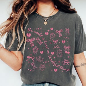 May include: Dark grey t-shirt featuring a pink graphic design with hearts, bows, and the text "the world is hard on beautiful things". The shirt has a relaxed fit, suitable for everyday wear.