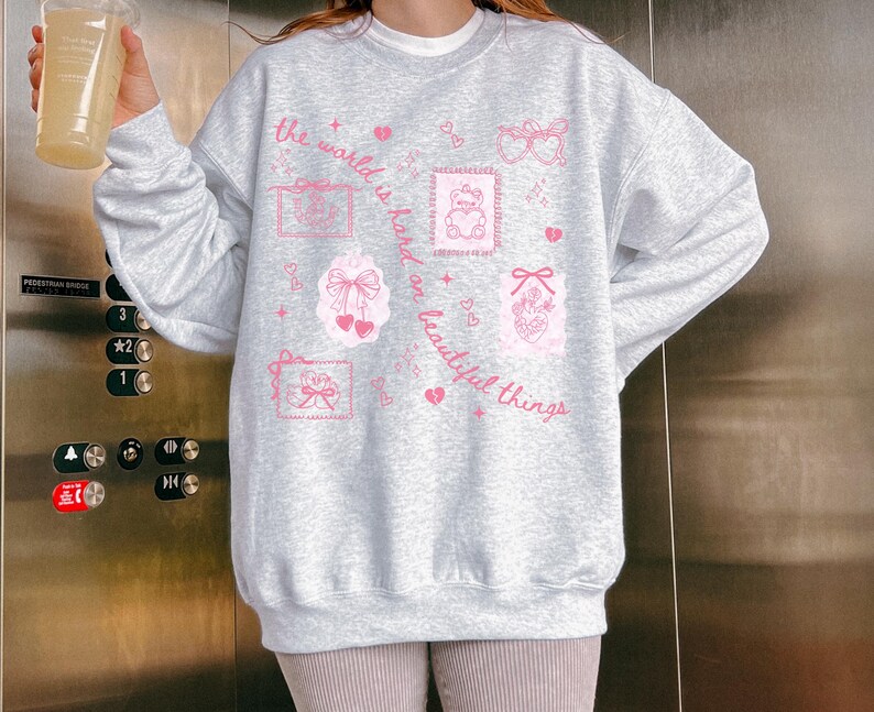 K&ouml;nnte beinhalten: Hellgraues Sweatshirt mit langen &Auml;rmeln und rosa Illustrationen von Schleifen, Teddyb&auml;ren und Herzen. Der Text "the world is hard on beautiful things" ist in rosa Schrift gedruckt. Das Sweatshirt hat einen Rundhalsausschnitt.