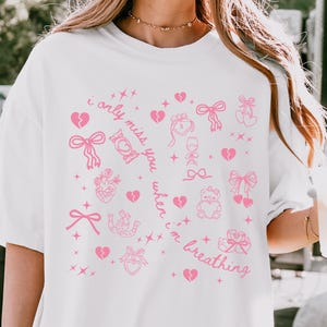 May include: White t-shirt with a pink graphic design featuring hearts, bows, and the text "i only miss you when i'm breathing". The design includes a variety of romantic symbols and text.