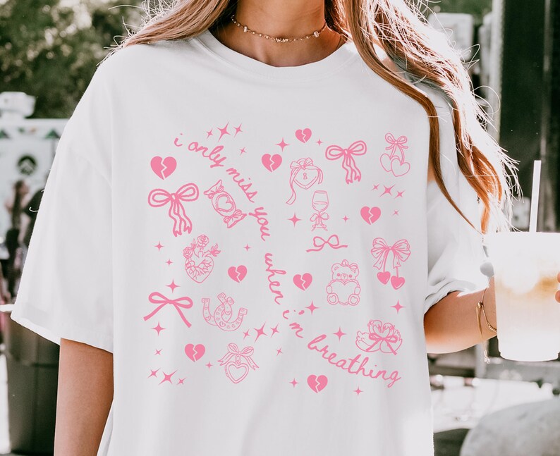 May include: White t-shirt with a pink graphic design featuring hearts, bows, and the text "i only miss you when i'm breathing". The design includes a variety of romantic symbols and text.