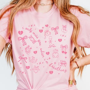 May include: Pink t-shirt with a repeating pattern of pink hearts, bows, and other romantic symbols. The text "i only miss you when i'm breathing" is printed on the shirt in a cursive font. The shirt is a light pink color.