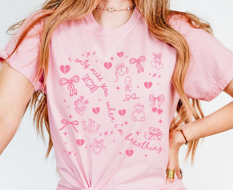 May include: Pink t-shirt with a repeating pattern of pink hearts, bows, and other romantic symbols. The text "i only miss you when i'm breathing" is printed on the shirt in a cursive font. The shirt is a light pink color.