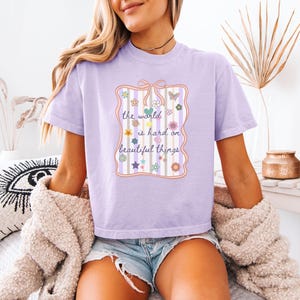 May include: A light purple t-shirt with the text "the world is hard on beautiful things" in a decorative frame with flowers and stars. The shirt has short sleeves and a crew neck.