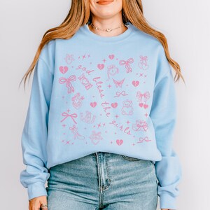 Może przedstawiać: Jasnoniebieska bluza z r&oacute;żowymi grafikami i napisem "God bless the girls". Projekt zawiera serca, kokardy, motyle i inne elementy dekoracyjne. Top z długim rękawem wykonany jest z miękkiego materiału.