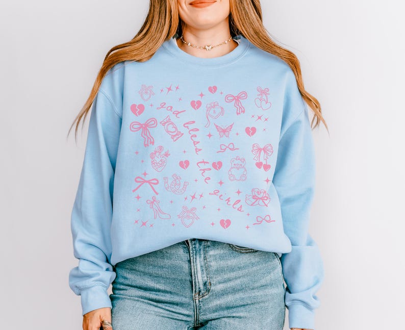 Może przedstawiać: Jasnoniebieska bluza z r&oacute;żowymi grafikami i napisem "God bless the girls". Projekt zawiera serca, kokardy, motyle i inne elementy dekoracyjne. Top z długim rękawem wykonany jest z miękkiego materiału.