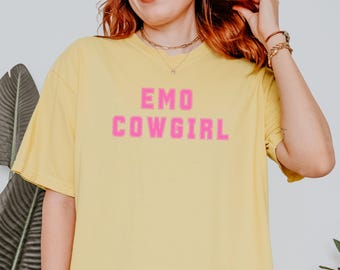 May include: A light yellow t-shirt with the words "EMO COWGIRL" in pink block letters. The shirt has short sleeves and a relaxed fit. The text is centered on the front of the shirt.