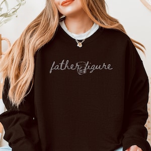 May include: A black sweatshirt featuring the phrase "father figure" in cursive writing. A football helmet graphic accompanies the word "figure". The sweatshirt is dark and has long sleeves.